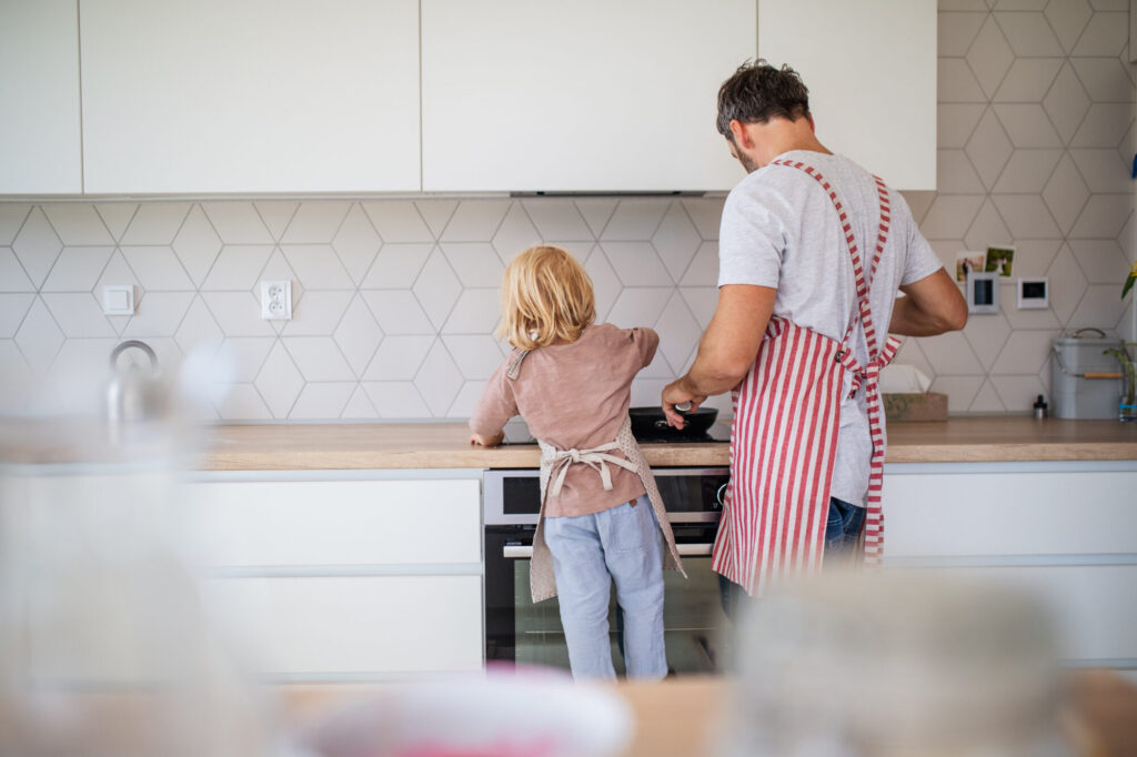 A rear view of small boy with father indoors in kitchen making pancakes.